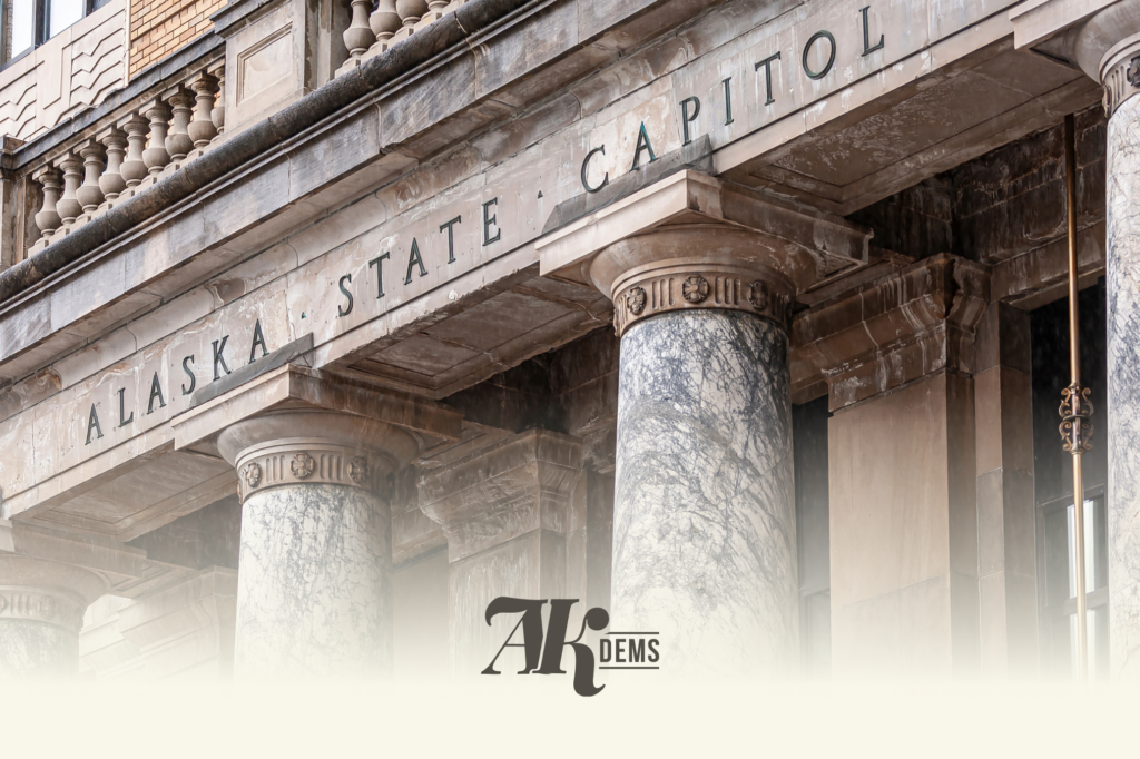 Federal-style building facade with carved columns and the words 'ALASKA STATE CAPITOL' carved along the entablature, stone and marble materials visible.