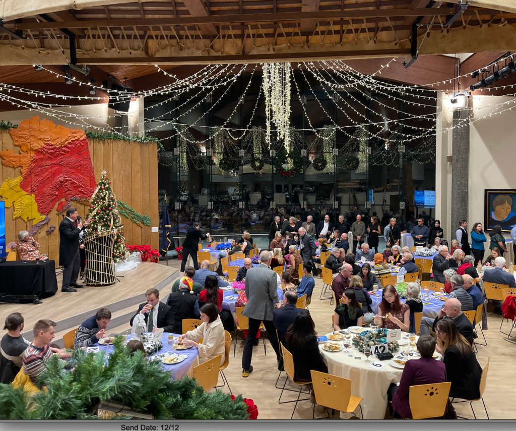 Banquet hall decorated for Christmas with hanging string lights, a speaker on stage, and many guests seated at round tables dining.