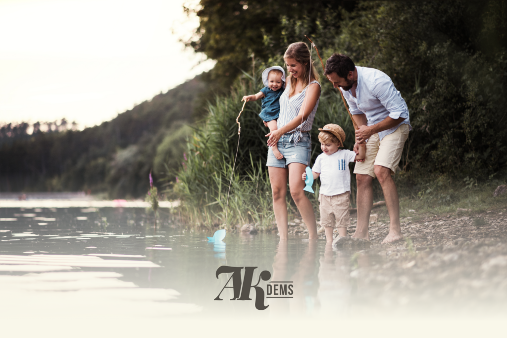 Family of four stands at the lakebank as parents help two young kids play with a paper boat by shallow water.