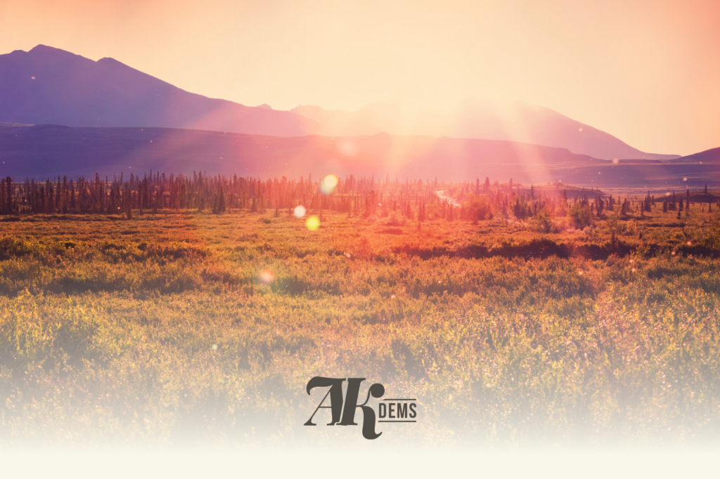 Sunlit mountain meadow with a distant range and trees, warm orange-pink hues and lens flare, logo at bottom center