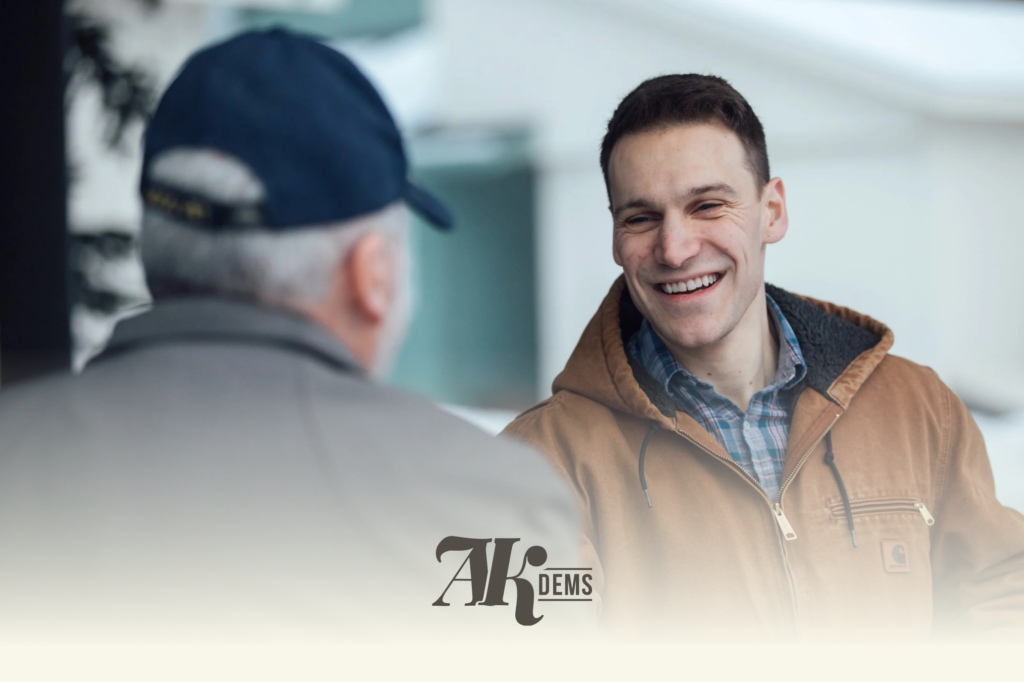 Two men sharing a friendly conversation; the younger man in a brown jacket smiles as he speaks to an older man wearing a dark cap (foreground blurred).