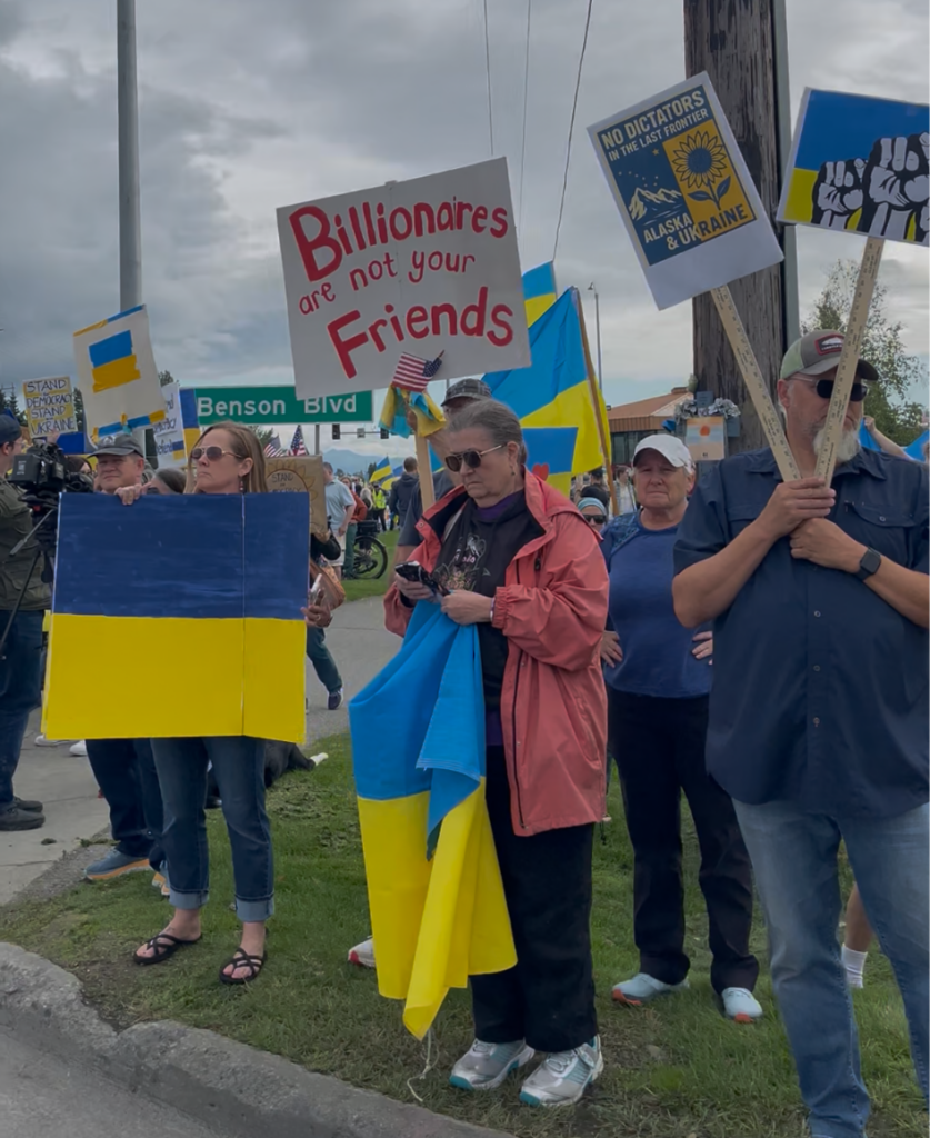 Protesters hold Ukrainian flags and signs, including a white sign saying 'Billionaires are not your Friends' on a street corner.