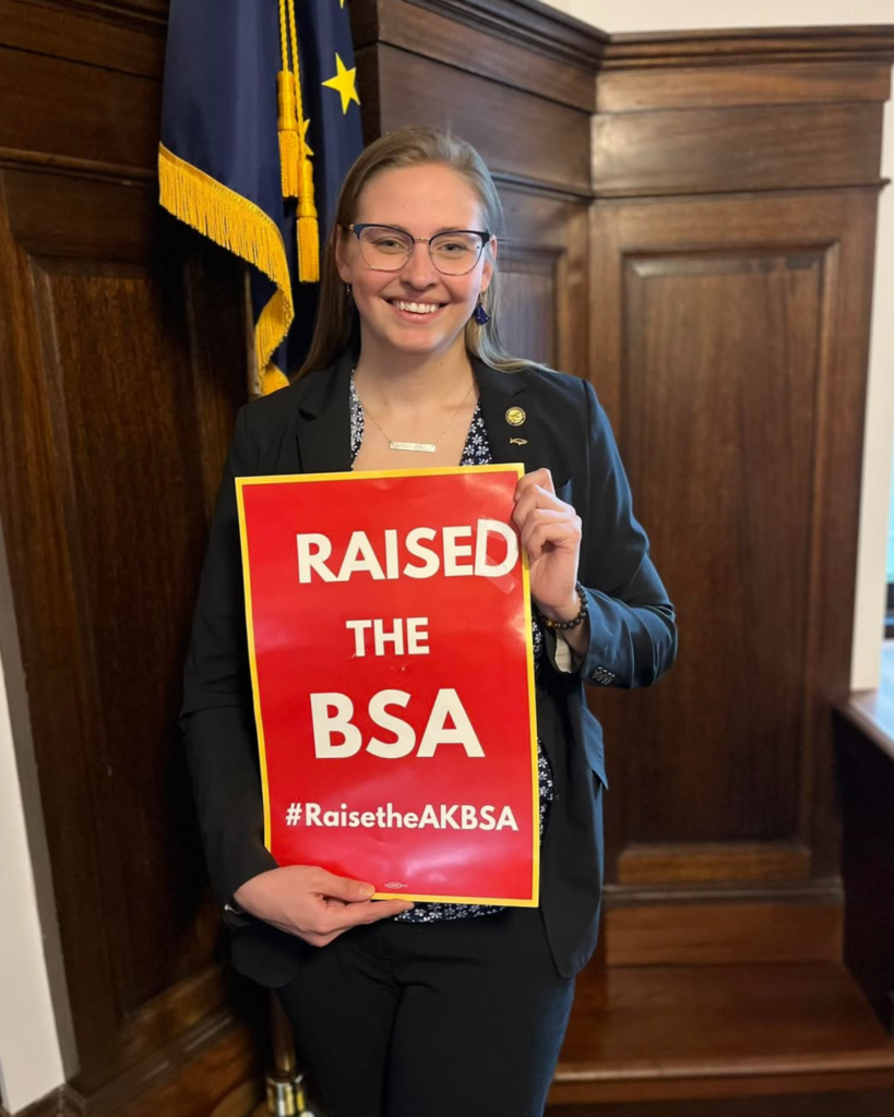 Smiling woman in a black blazer holds a red poster reading 'RAISED THE BSA' with the hashtag #RaiseTheAKBSA, standing in a wood-paneled room with a flag behind her.