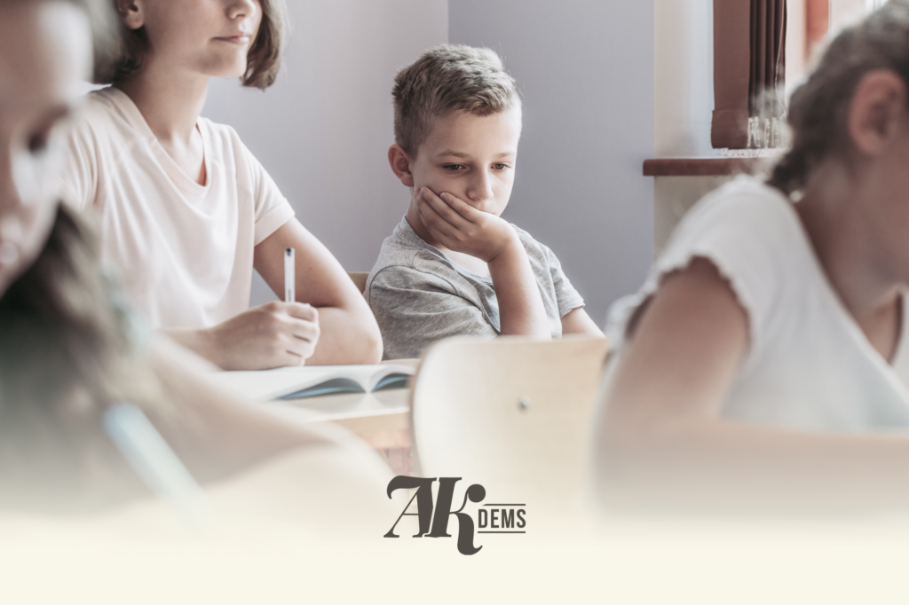 Young students sit at a classroom table; a boy in a gray shirt rests his chin on his hand, deep in thought, while others work nearby.