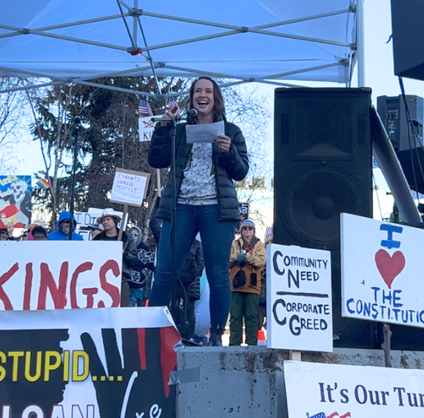 Woman speaking into a microphone on a protest stage, addressing a crowd with signs about community needs and corporate greed in the background.