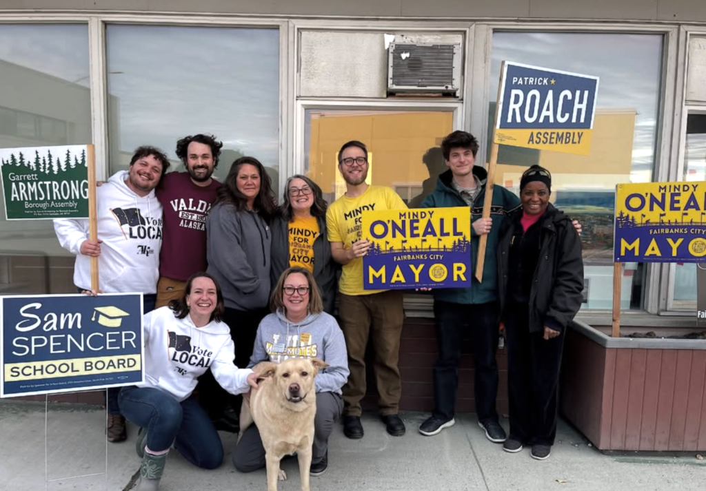 Group of diverse volunteers posing outdoors with campaign signs for Mindy Oneall mayor, Patrick Roach assembly, and Sam Spencer school board; a dog sits front center.