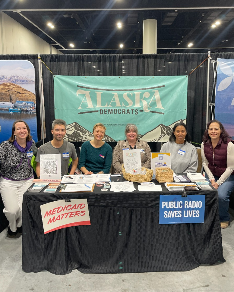 Six people sit or stand behind a long table at an Alaska Democrats booth, with a teal banner reading ‘ALASKA DEMOCRATS’ and signs saying ‘Medicaid MATTERS’ and ‘PUBLIC RADIO SAVES LIVES’. Pamphlets, QR code, and baskets are on the table.
