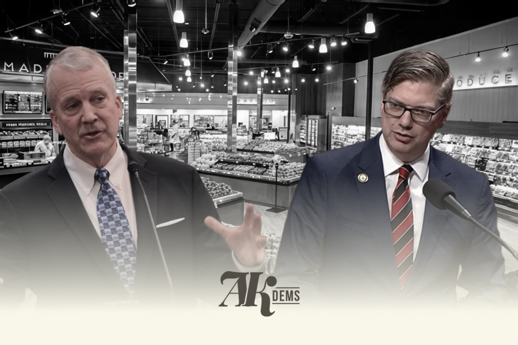 Two men in suits speak at microphones in a grocery store setting with produce displays behind them.