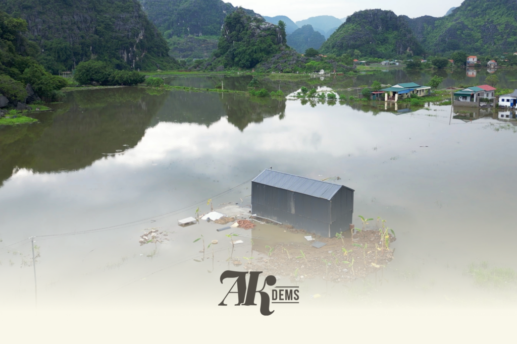 Flooded rural area with submerged fields and houses, green hills and rocky mountains in the distance.