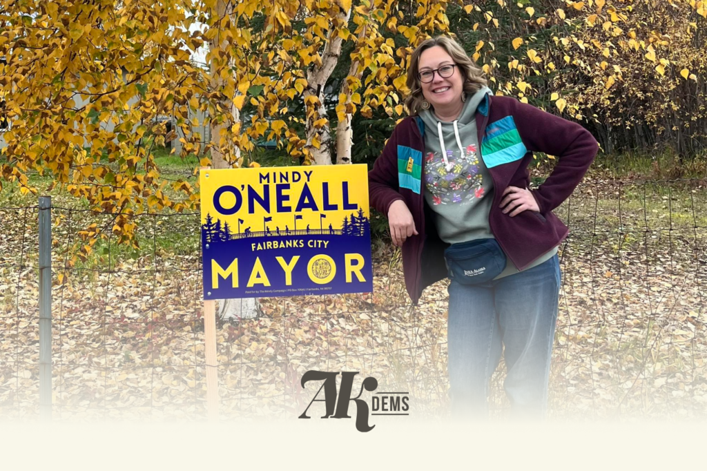 Smiling woman in glasses stands beside a bright yellow campaign sign that reads 'MINDY O'NEALL MAYOR' among autumn leaves and trees.
