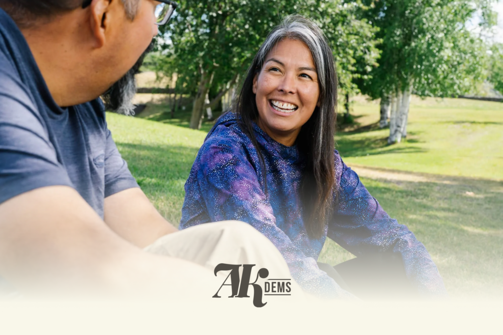 Two people outdoors in a park, smiling at each other as they chat, with greenery in the background and a logo at the bottom edge.