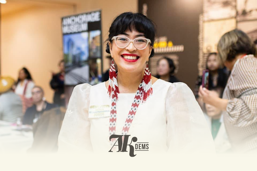 Smiling woman with white-framed glasses and red-and-white beaded earrings at a crowded event; badge visible on her blouse.