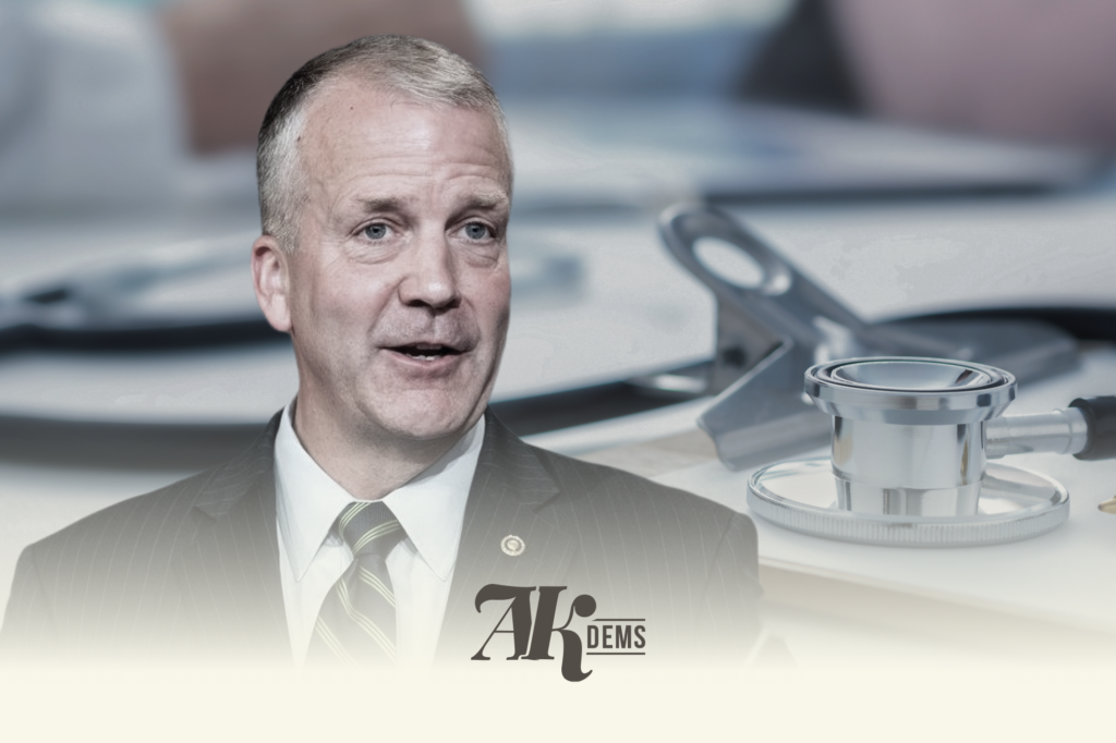 Portrait of a man in a suit speaking, with blurred medical equipment in the background and an AK DEMS logo at the bottom right.