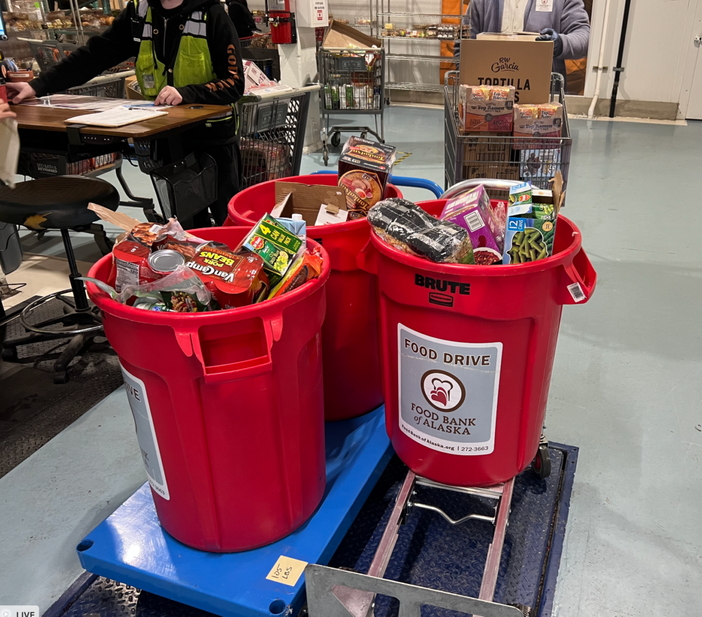 Red Food Drive bins brimming with canned goods and boxes at a grocery store; Food Bank of Alaska label visible.