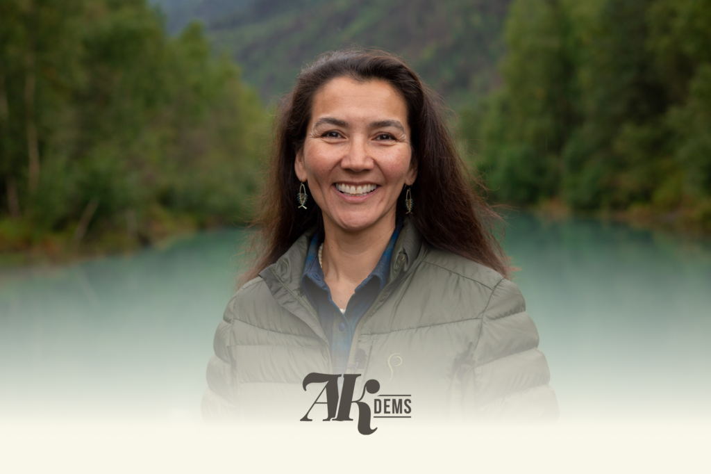 Smiling woman in a green quilted jacket outdoors with a forested lake backdrop; logo 'AK DEMS' partially visible over her torso.