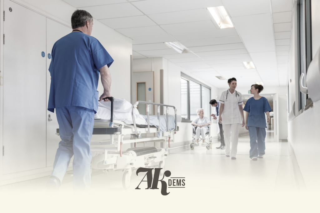 Medical staff in blue scrubs push a patient on a wheeled stretcher down a bright hospital corridor, with a nurse and another caregiver in the background.