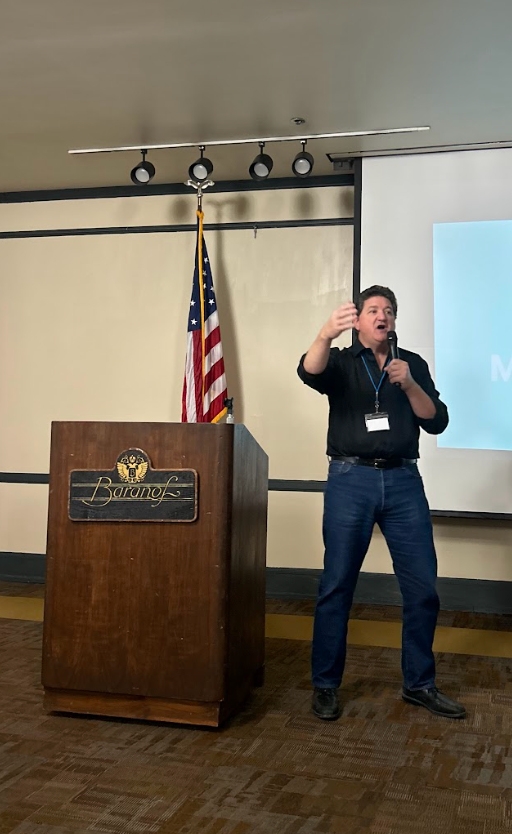 Male presenter in a black shirt and jeans speaks at a wooden podium with an American flag behind him and a projection screen to his right in a conference room.