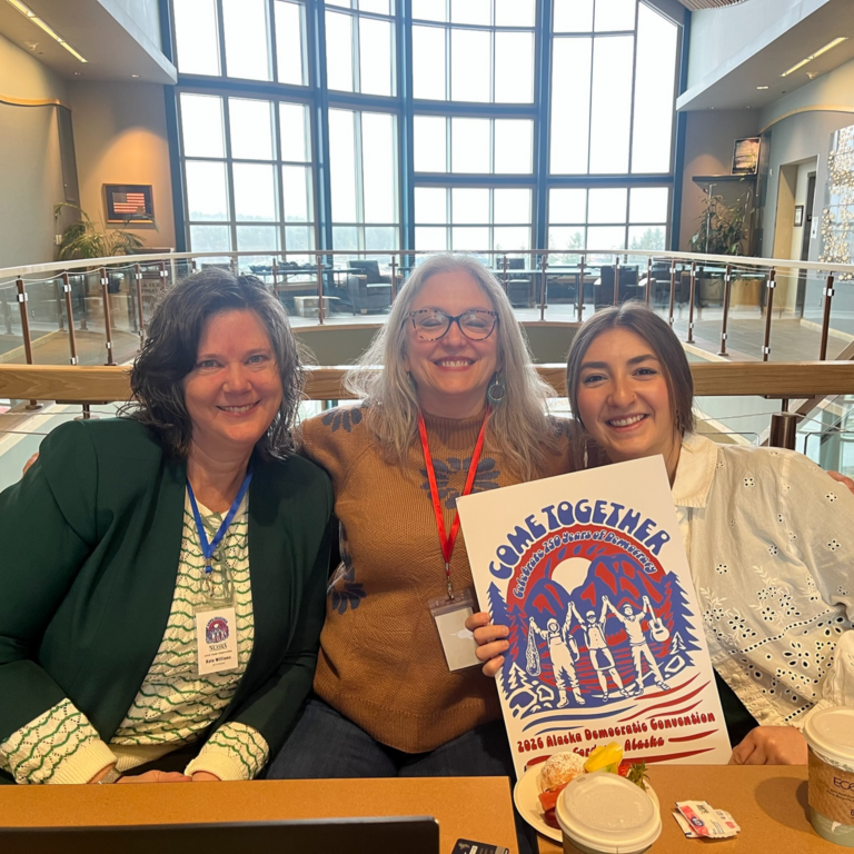 Three women smiling in a bright atrium, holding a poster that reads 'Come Together' for the 2026 Alaska Democratic Convention while wearing lanyards.