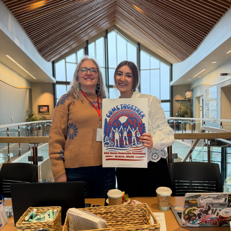 Two smiling women stand in a bright atrium, holding a colorful poster that reads 'Come Together' with a group illustration behind them.
