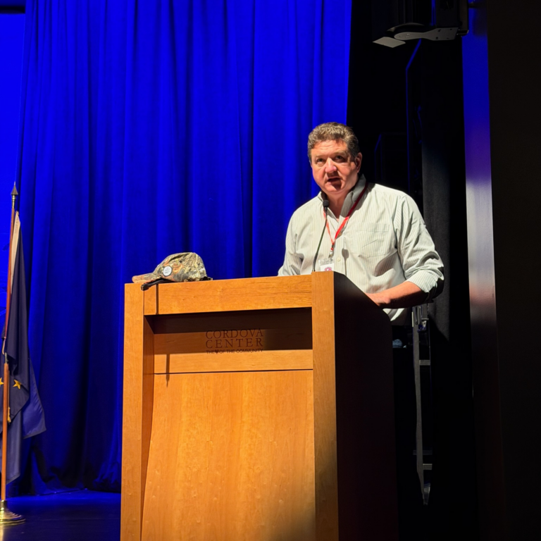 Man speaking at a wooden podium on a stage with blue curtains; a camouflage cap rests on the podium.