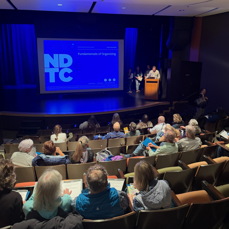 Speaker at a wooden podium on a stage, presenting to an audience in a dark theater with a blue NDTC slide projected on screen reading 'Fundamentals of Organizing'.