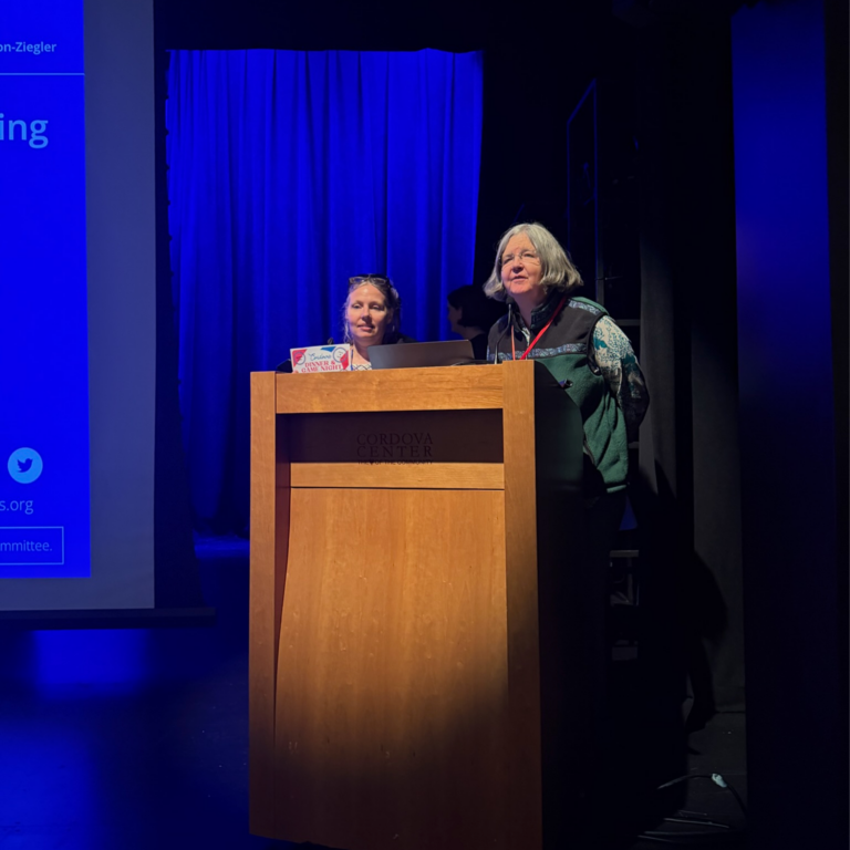 Two women stand at a wooden lectern on a blue-lit stage; one speaks into a microphone while the other watches with a laptop.
