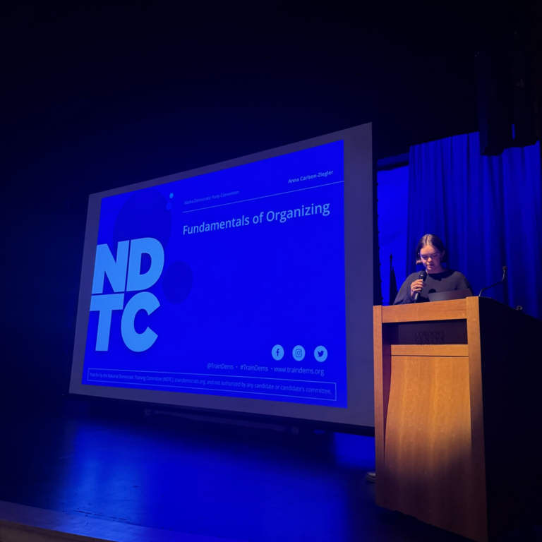 Speaker at a wooden podium presenting a slide titled 'Fundamentals of Organizing' on a large blue screen in a dark auditorium, with bold 'NDTC' graphics.