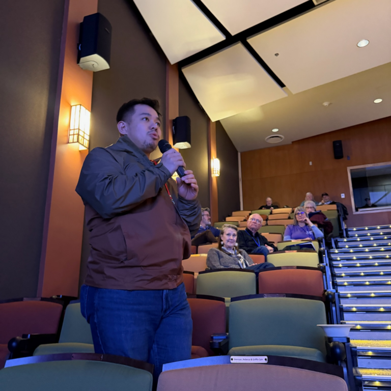 Man stands in a dim auditorium, speaking into a handheld microphone during a lecture or Q&A. Audience seated behind him.