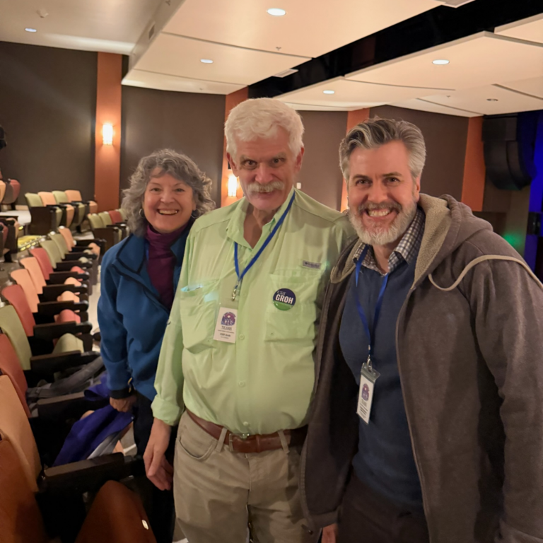 Three smiling adults pose together in an auditorium with colorful seats in the background, wearing name badges for a conference or event.