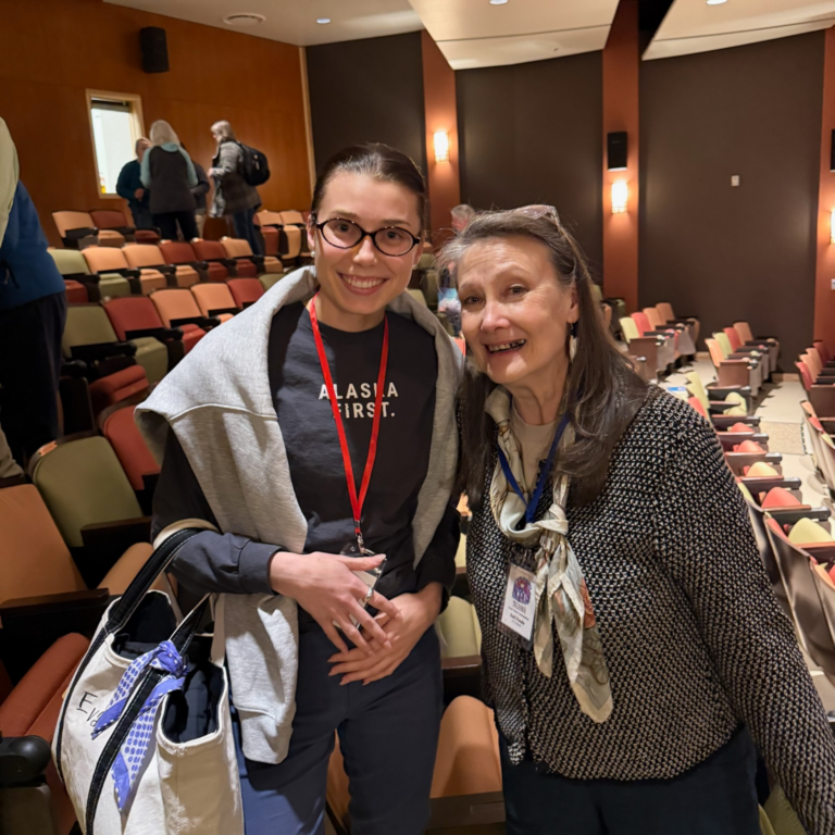 Two smiling women pose together in a theater auditorium with colorful, empty seats in the background; one wears glasses and a red lanyard, the other has a scarf and an event badge.