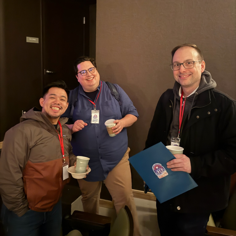 Three men pose together at a casual event, each holding a coffee cup and wearing lanyard badges.