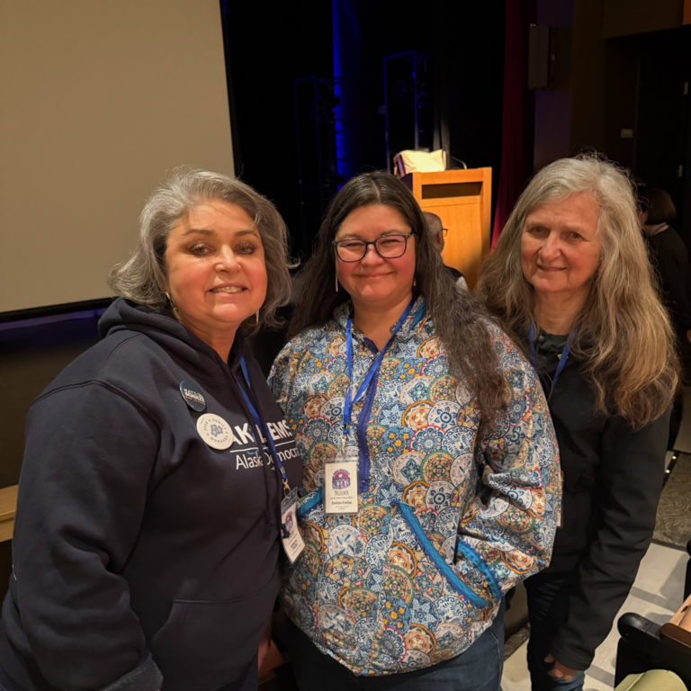 Three smiling women posing together at an event, each wearing a lanyard and name badge in a conference setting.