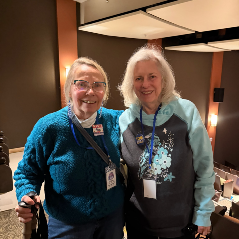 Two smiling older women pose together in a theater lobby with lanyards and badges; one in a teal fleece, the other in a turquoise hoodie with floral design.