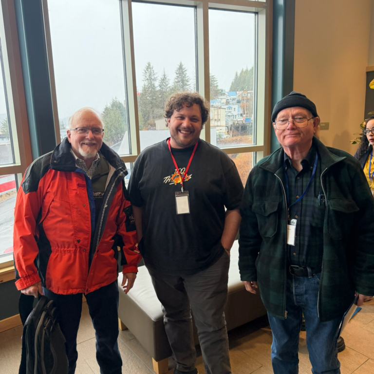 Three men stand indoors by a large window, smiling for the camera. Left man in an orange jacket with glasses, middle man in a black T-shirt with a red lanyard, right man in a dark green jacket and beanie.