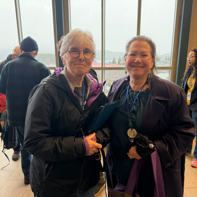 Two smiling women stand together indoors at a conference, one with white hair and glasses holding a blue folder neglected by her side.