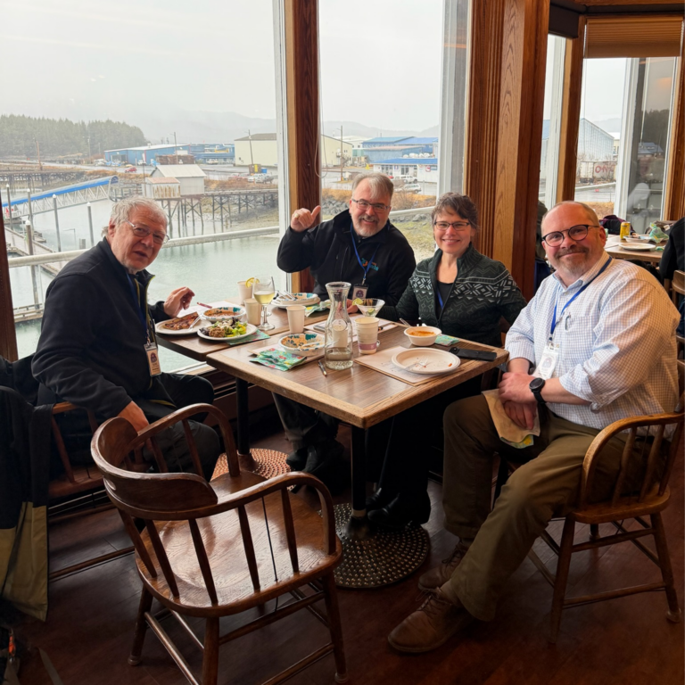 Four adults seated around a wooden table eating lunch by large windows with a harbor view outside.