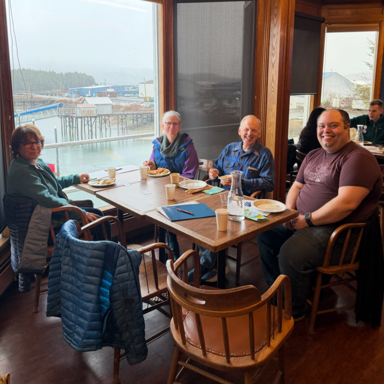 Four people dining at a waterfront restaurant table, smiling and enjoying a meal by large windows overlooking the dock area.