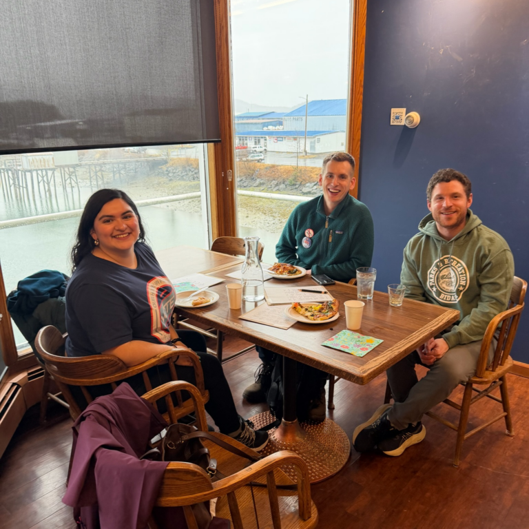 Three friends sit around a wooden table enjoying a meal by a large window, smiling at the camera with plates and drinks in front of them.