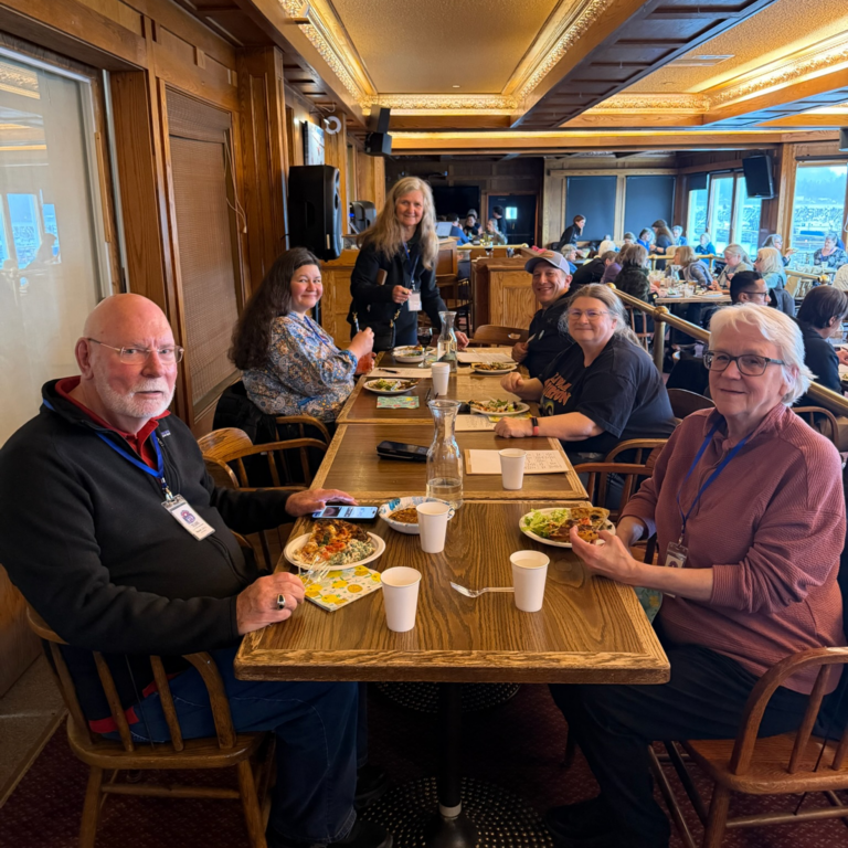 Group of seniors dining at a long wooden table in a busy restaurant, plates of food and drinks in front of them, smiling at the camera during a meal event.