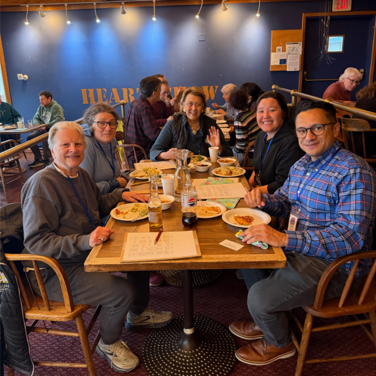 Six adults sit around a wooden table in a casual restaurant, smiling at the camera amid plates of food, drinks, and a pitcher; they wear lanyards and name badges, with a blue wall and warm lighting in the background.