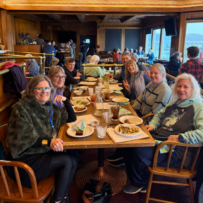 Eight people sit around a long table in a cozy wood-paneled restaurant, enjoying plated meals and smiles at the camera.
