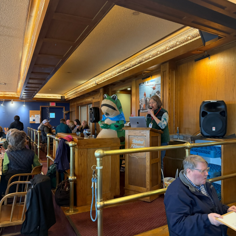 Person speaking at a wooden podium with a microphone; a frog mascot stands beside them in a dining hall setting.
