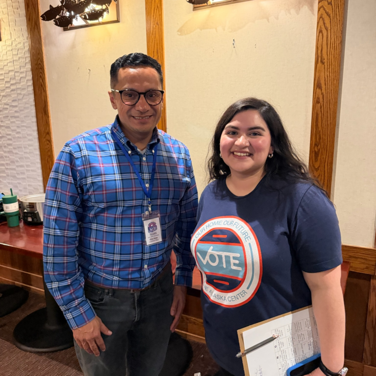 Two smiling people posing indoors: a man in a blue plaid shirt with a name badge and a woman in a navy Vote shirt holding a clipboard.