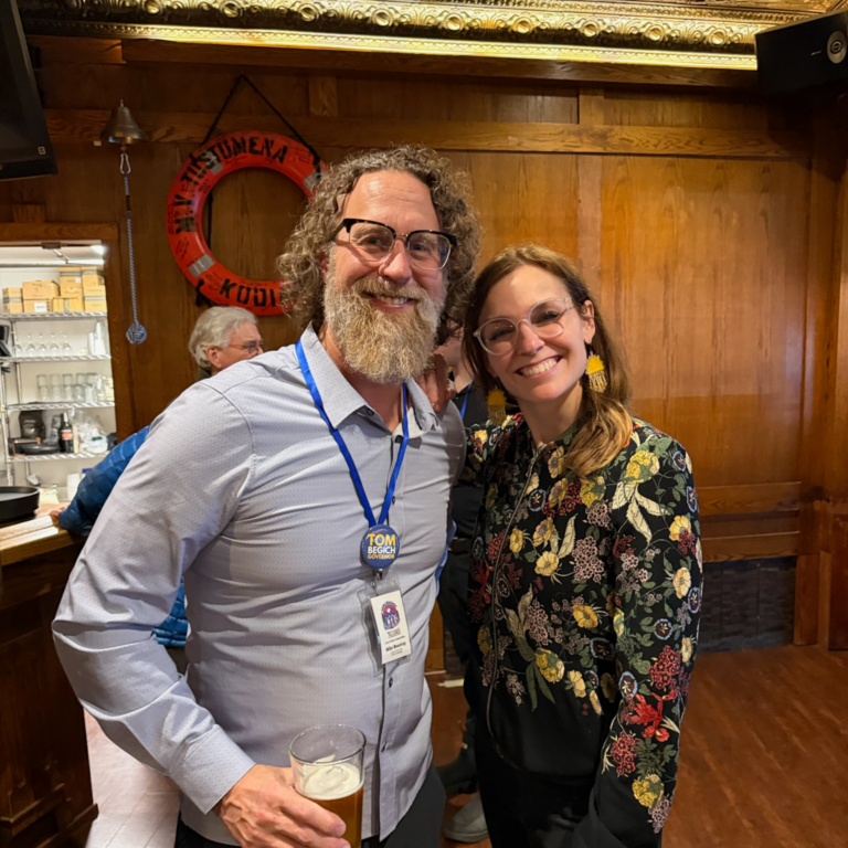 Smiling man with gray beard and glasses in a blue shirt holding a beer, standing next to a smiling woman in a floral top with yellow earrings in a wood-paneled room.