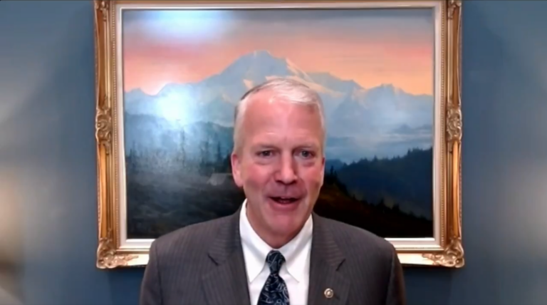 Smiling man in a striped suit speaks during a video call with a mountain landscape painting behind him on a blue wall.