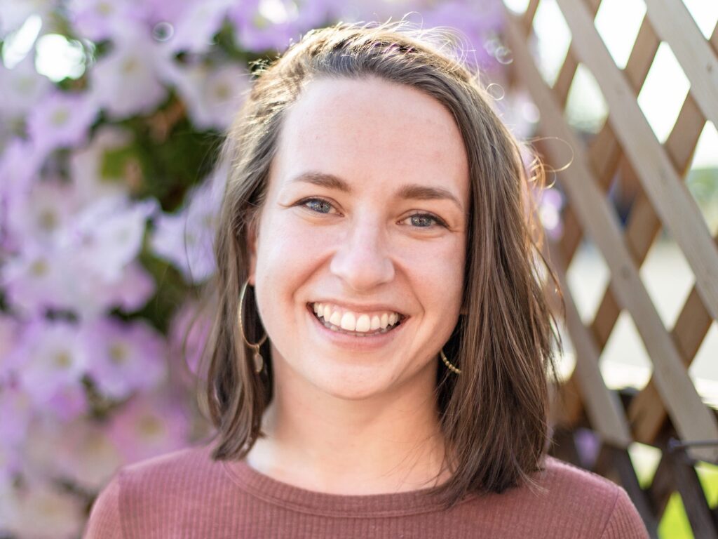 Close-up of a smiling woman with shoulder-length brown hair in a maroon sweater, with purple flowers and a lattice fence in the background.