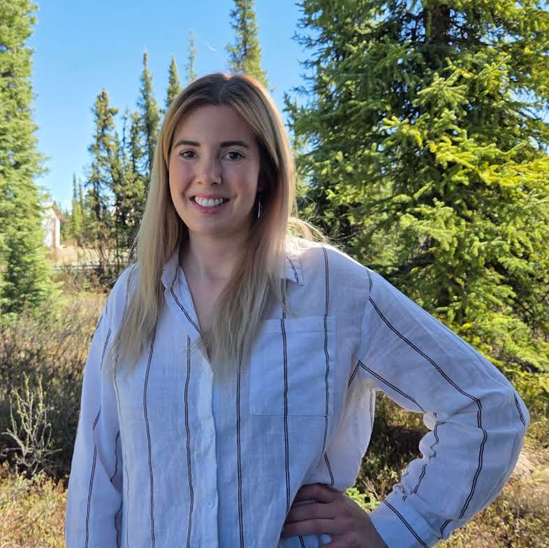 Smiling woman with long blonde hair standing outdoors among evergreen trees, wearing a white striped button-down shirt.