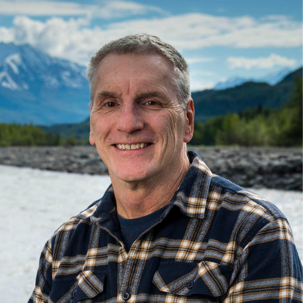 Smiling man in a blue and tan plaid shirt posing outdoors by a rocky river with mountains and blue skies in the background.