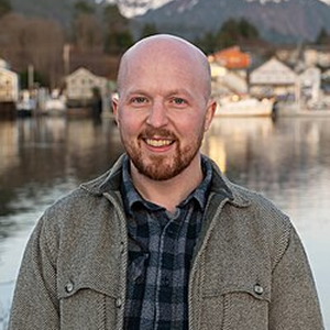 Smiling, bald man with a beard wearing a gray jacket and plaid shirt, standing by a marina with boats and a town in the background.