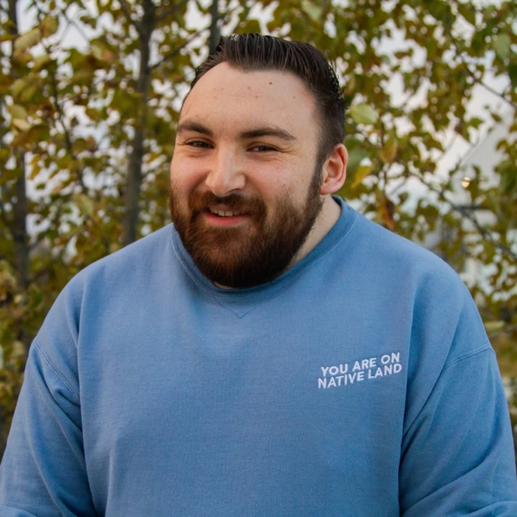 Smiling man with a beard in a blue sweatshirt reading 'YOU ARE ON NATIVE LAND', outdoors with fall leaves behind him.